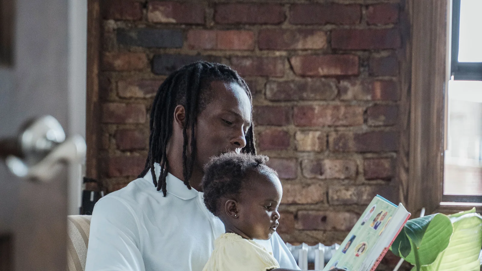 Father holding a young child on his lap while sharing an illustrated book in an armchair.