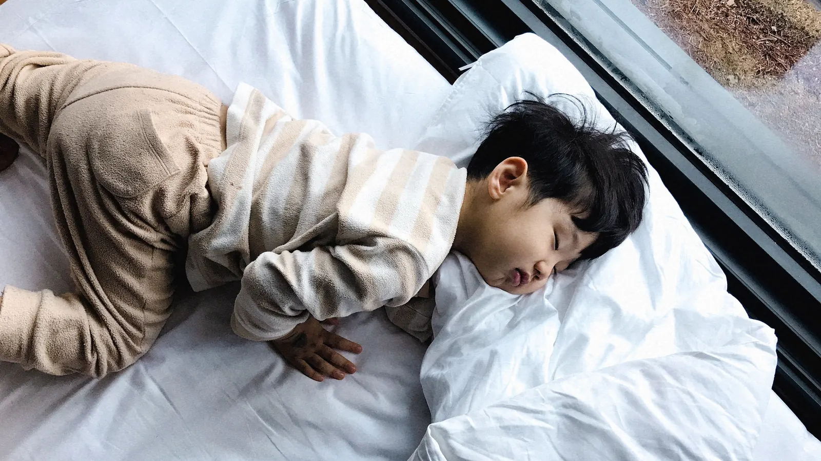 Child sleeping on white bedding beside a window.